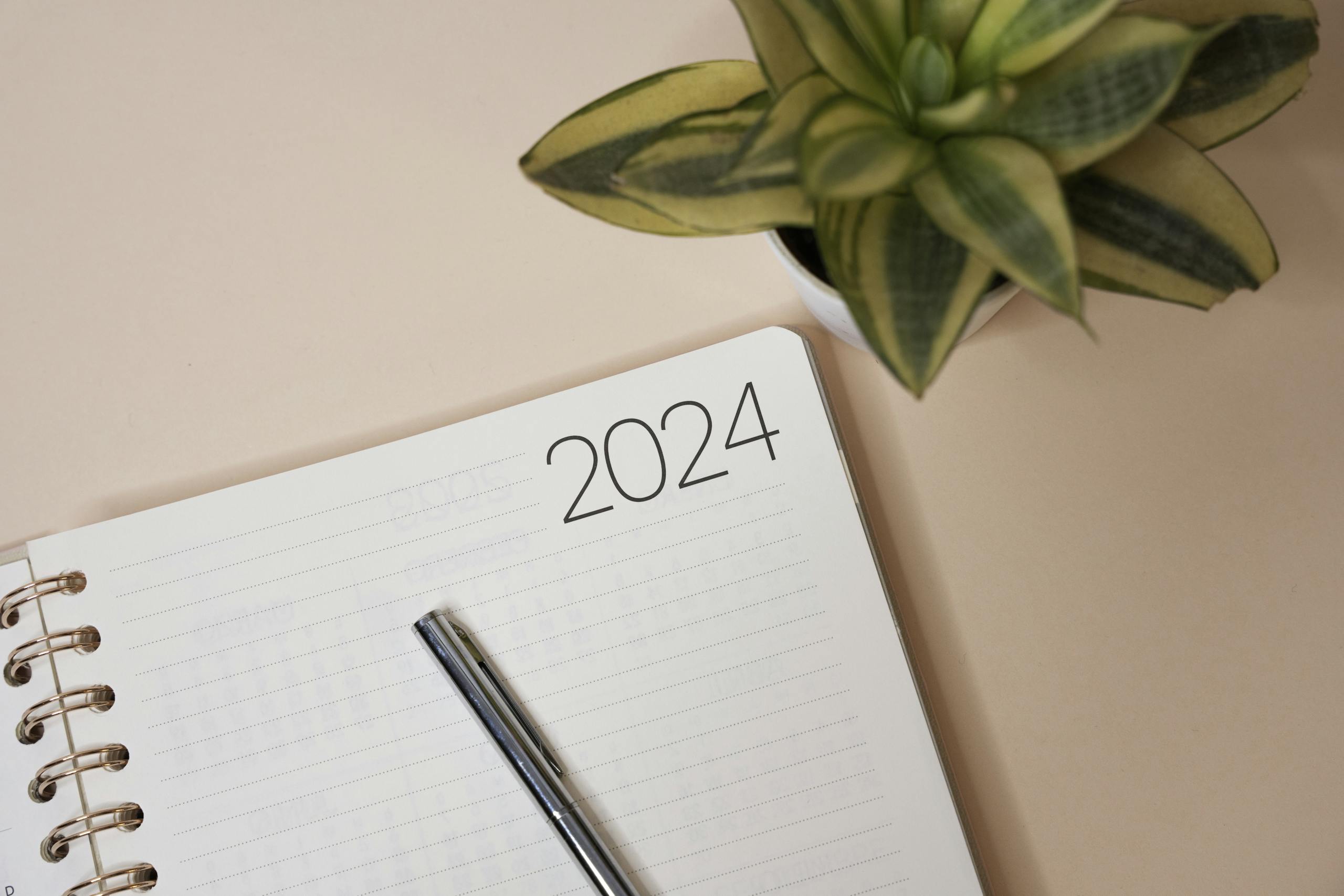 A simple 2024 planner with a pen on a peach background beside a potted plant, perfect for organization and planning.