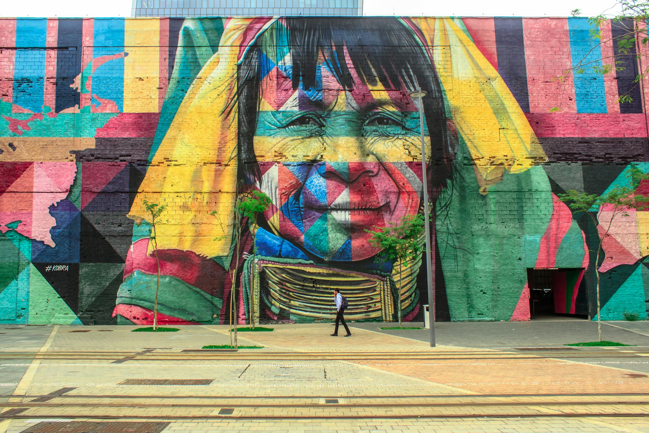 Colorful mural depicting a native face on an urban street wall with a passerby.